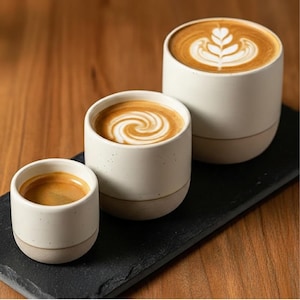 May include: Three ceramic coffee cups with latte art. The cups are white with a tan base and filled with coffee. The coffee art includes a swirl and a floral design. The cups are arranged on a black rectangular tray.
