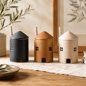May include: Three ceramic house-shaped containers with lids and small spoons. The containers are in black, orange, and cream colours. Each has square window cutouts and a door. The containers are on a wooden table.