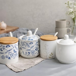 May include: Four white ceramic kitchen canisters with lids and spoons. Two canisters have blue floral patterns, one has a leaf design, and one is plain. The lids are made of bamboo.