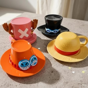Puede incluir: Cuatro tazas de café y platillos de cerámica en varios colores y diseños. Una taza rosa presenta una cruz blanca y cuernos marrones. Una taza naranja tiene diseños de caras azules y blancas. Una taza amarilla tiene forma de sombrero de paja con una banda roja. Una taza negra tiene un diseño azul y blanco.