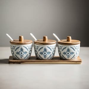 May include: A set of three ceramic spice jars with wooden lids and small spoons, resting on a wooden tray. The jars have a white base with a blue floral pattern. The lids and tray are made of natural wood.