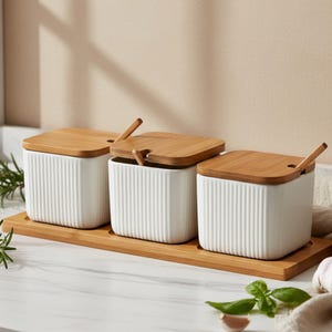 Puede incluir: Juego de tres botes de cerámica blanca para especias con tapas y cucharas de bambú, colocados sobre una bandeja de bambú. Los botes tienen un diseño acanalado. Las tapas tienen una pequeña muesca para las cucharas.