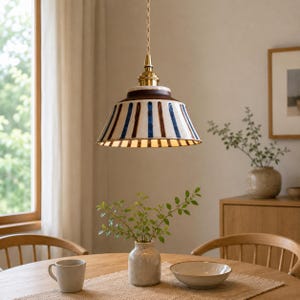 Pode incluir: Uma luminária pendente de cerâmica com uma cúpula alargada apresentando listras verticais azuis e marrons sobre um fundo creme, suspensa por um cabo de tecido trançado a partir de um suporte de latão polido fixado ao teto.
