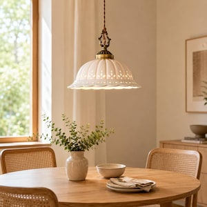 May include: A white ceramic pendant light with a scalloped edge and intricate cut-out patterns, suspended by a twisted brown cord from an ornate brass fixture. The dome-shaped shade emits a soft glow, featuring a fluted design with small holes.