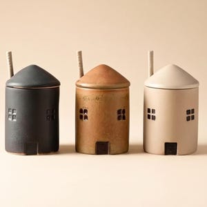 Puede incluir: Tres tarros de cerámica con forma de casa en negro, marrón y crema. Cada tarro tiene una tapa cónica, ventanas cuadradas y una pequeña puerta. Una pequeña cuchara descansa sobre cada tapa. Estos tarros decorativos son ideales para especias.