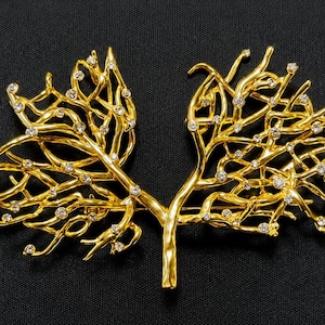 May include: A gold-toned brooch shaped like two branching coral structures joined at the base. The metallic surface features a textured, organic finish and is accented with numerous small, clear, round rhinestones embedded throughout the branches.