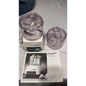 May include: A white Cuisinart Mini-Prep Processor with a clear plastic work bowl, a separate clear lid, and a power cord. The base features two gray buttons labeled LO and HI. An instruction and recipe booklet for the DLC-1 model is included.