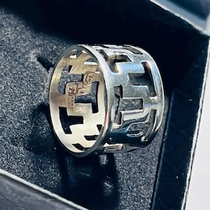 Könnte beinhalten: Ein breiter Silberring mit einem sich wiederholenden geometrischen Ausschnittmuster. Die Innenseite des Rings ist deutlich mit dem Stempel 925 versehen, was auf eine Zusammensetzung aus Sterlingsilber hinweist. Der Ring liegt in einer dunklen Box.