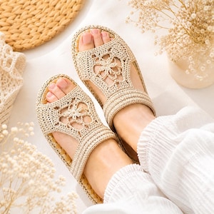 Op de afbeelding: Een paar beige gehaakte sandalen met een open teenontwerp en een centraal bloemmotief op de bovenste band. De sandalen hebben een geweven textuur en een stevige zool, wat zorgt voor een neutrale uitstraling die past bij de lichte stof.