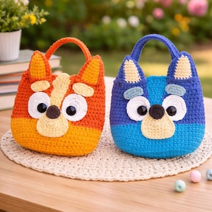 Könnte beinhalten: Zwei handgefertigte Häkeltaschen, eine orange und gelb, die andere blau und türkis. Jede Tasche ist wie ein Cartoon-Hundekopf geformt, mit großen Augen, einer Nase und Ohren. Sie haben einen Griff oben und sitzen auf einer beigen Tischmatte.