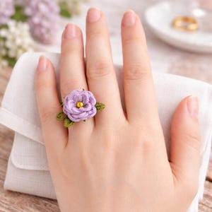 May include: A ring featuring a crocheted lavender flower with a small yellow bead at its centre, accented by two dark green crocheted leaves. The floral design is crafted with intricate thread work, showcasing a layered petal structure.