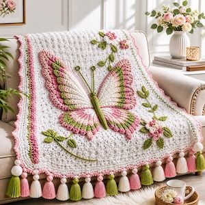 Op de afbeelding: Een witte gehaakte deken met een groot centraal vlindermotief in roze en groene tinten. De deken is versierd met driedimensionale gehaakte bloemen en groene ranken, afgewerkt met een schulprand en roze, groene en witte kwastjes.