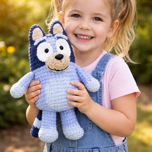 Op de afbeelding: Een gehaakt blauw hondenkussen met een lichtbruine snuit en zwarte ogen. De knuffel wordt vastgehouden door een jong kind dat een spijkerbroek met tuinbroek en een roze shirt draagt. De achtergrond is een wazige buitenscène met zonlicht.