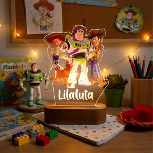 May include: A decorative night light with characters from Toy Story, including Buzz Lightyear and Woody. The base is made of wood, and the name "Lilaluta" is displayed in white. The lamp is on a wooden table with other toys and a notebook.