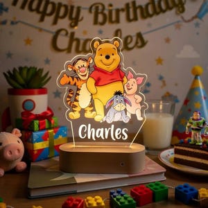 Könnte beinhalten: Ein Nachtlicht im Winnie-the-Pooh-Design mit den Figuren Tiger, Winnie Puuh, Ferkel und I-Aah. Der Name "Charles" ist unter den Figuren zu sehen. Das Licht steht auf einem Holzsockel, mit Geburtstagsdekorationen im Hintergrund.