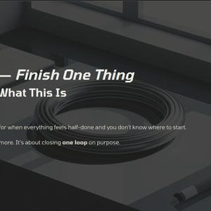 May include: A dark, grayscale image featuring text and objects. The text reads "PDF v2 - Finish One Thing" and "Page 1 - What This Is." A coiled, dark gray object and a pen are visible. The image is on a desk.