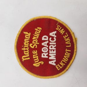 Puede incluir: Un parche bordado circular con fondo rojo y borde cosido en amarillo. El texto dice National June Sprints ROAD AMERICA ELKHART LAKE, WIS. en hilo amarillo y blanco, dispuesto en un diseño curvo a través del centro.