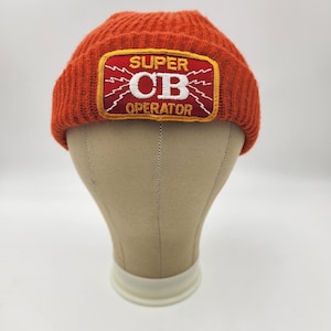 Könnte beinhalten: Eine orangefarbene Strickmütze mit einem rechteckigen Aufnäher. Der Aufnäher hat einen roten Rand und trägt die Worte "SUPER CB OPERATOR" in Weiß und Gelb. Die Mütze wird auf einem Schaufensterkopf präsentiert.