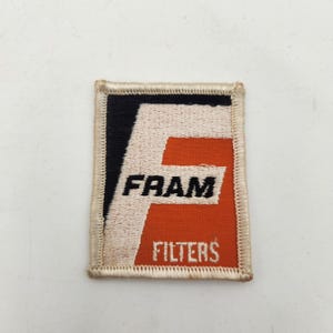 May include: A rectangular embroidered patch featuring the Fram logo. The design includes a large white letter F on a black and orange background. The word FRAM is stitched in black, and the word FILTERS is stitched in white. The patch has a cream border.
