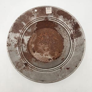 May include: A circular metal hubcap featuring a silver-toned finish with significant brown rust accumulation in the center and along the outer rim. A small white rectangular sticker with the text "RUBY CITY 44 E. Main Franklin, N.C. #2.95" is attached.