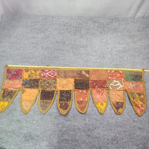 May include: A rectangular fabric valance featuring a patchwork of multi-colored embroidered textile squares with metallic thread accents and sequins. The bottom edge has eight pointed pennant-style drops. A yellow measuring tape shows a width of 58 inches.