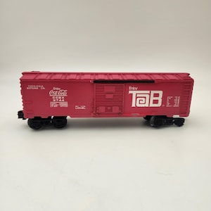 May include: A red toy train car with black wheels. The car features the Coca-Cola and Tab logos in white. The text "Enjoy Coca-Cola" and "Enjoy Tab" are visible on the side of the car. The car is a model of a freight train car.