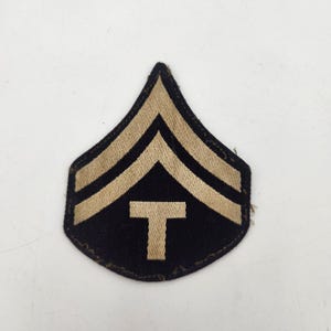 以下が含まれることがあります： 黒い背景に上向きのベージュ色のシェブロンストライプが2本入った、盾形のミリタリーランクパッチ。シェブロンの下には、黒い生地の中央にベージュ色の文字「T」が配置されています。パッチにはステッチが施された縁と尖った上部があります。