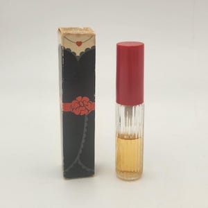 May include: A tall, rectangular cardboard box featuring a black dress illustration with a red waist flower and a small red heart, standing next to a clear, vertically ribbed glass perfume bottle with a solid red cylindrical cap and amber liquid.