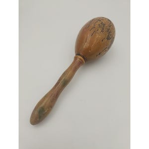 May include: A vintage wooden maraca with a light brown, oval-shaped head and a long, slender handle. The head shows signs of wear, with some of the paint chipped off. The handle has a lighter brown color with some green accents.