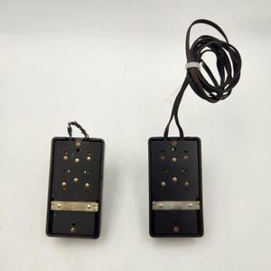 May include: Two rectangular black electronic components with metallic contact points and screws. One unit features a frayed black wire connection at the top, while the other has a long, coiled black electrical cord secured with a white plastic tie.