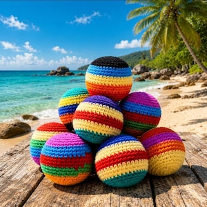 May include: A collection of seven colorful, crocheted balls arranged on a wooden surface. Each ball features horizontal stripes in a rainbow of colors, including red, orange, yellow, green, blue, purple, and black. The balls are set against a beach backdrop.