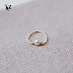 K&ouml;nnte beinhalten: Ein zarter Ring aus kleinen wei&szlig;en Perlen mit einer einzigen gro&szlig;en wei&szlig;en Perle in der Mitte. Der Ring ist mit zwei kleinen goldenen Perlen auf beiden Seiten der Perle verziert.