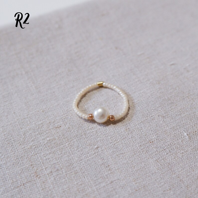 K&ouml;nnte beinhalten: Ein zarter Ring aus kleinen wei&szlig;en Perlen mit einer einzigen gro&szlig;en wei&szlig;en Perle in der Mitte. Der Ring ist mit zwei kleinen goldenen Perlen auf beiden Seiten der Perle verziert.