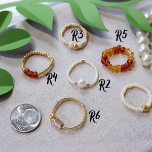 K&ouml;nnte beinhalten: Sechs Perlenringe in verschiedenen Farben und Stilen. Die Ringe sind mit R1 bis R6 beschriftet. Die Ringe liegen auf einer beigefarbenen Stoffoberfl&auml;che.