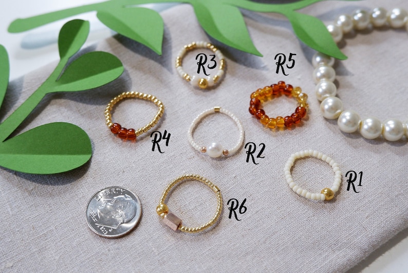 K&ouml;nnte beinhalten: Sechs Perlenringe in verschiedenen Farben und Stilen. Die Ringe sind mit R1 bis R6 beschriftet. Die Ringe liegen auf einer beigefarbenen Stoffoberfl&auml;che.