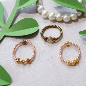 May include: Three beaded rings with different colored beads. The rings are on a light brown fabric background with green paper leaves.