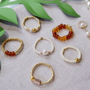 K&ouml;nnte beinhalten: Eine Sammlung von sechs Perlenringen in verschiedenen Farben und Stilen. Die Ringe sind aus kleinen Perlen in Wei&szlig;, Braun und Gold gefertigt. Einige Ringe haben einen Perlmutt- oder Goldakzent.