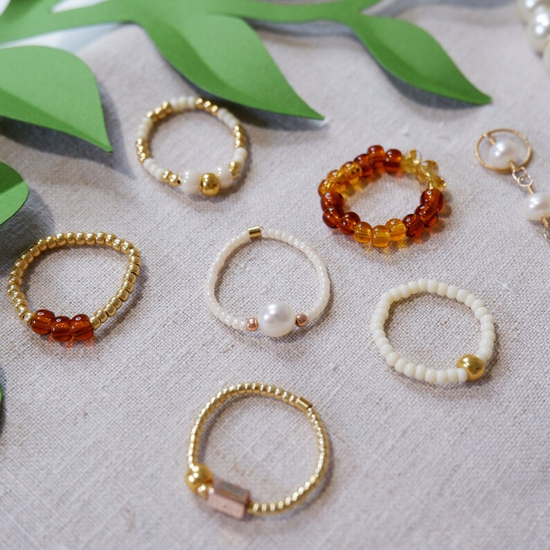 K&ouml;nnte beinhalten: Eine Sammlung von sechs Perlenringen in verschiedenen Farben und Stilen. Die Ringe sind aus kleinen Perlen in Wei&szlig;, Braun und Gold gefertigt. Einige Ringe haben einen Perlmutt- oder Goldakzent.