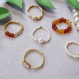 May include: Six beaded rings with different colors and materials. The rings are made of gold, white, and amber beads. Some rings have a pearl or a gold accent.