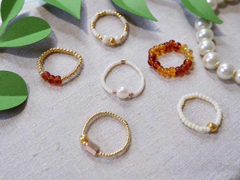 K&ouml;nnte beinhalten: Sechs Perlenringe in verschiedenen Farben und Materialien. Die Ringe bestehen aus goldenen, wei&szlig;en und bernsteinfarbenen Perlen. Einige Ringe haben einen Perlen- oder Goldakzent.