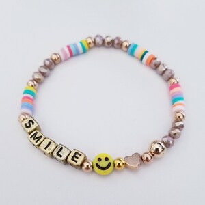 May include: A gold beaded bracelet with a rainbow striped charm, a smiley face charm, a heart charm, and gold letter beads spelling out "SMILE".