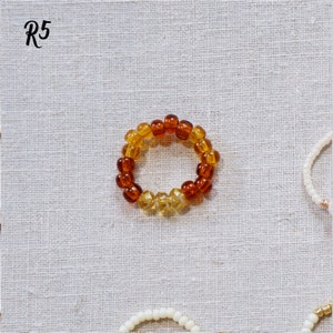 K&ouml;nnte beinhalten: Ein Ring aus Bernstein- und Goldperlen. Der Ring liegt auf einem wei&szlig;en Stoffhintergrund. R5 steht in der oberen linken Ecke des Bildes.