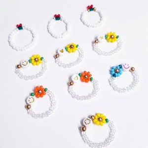 May include: Nine beaded rings with different flower and smiley face charms. The rings are white with gold accents and feature various colors, including yellow, orange, blue, and green.