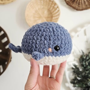 May include: A handmade blue and white crochet whale toy with a small tail and flipper. The whale has a black eye and a small pink cheek. The toy is held in a hand.
