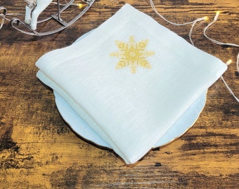 May include: A white linen napkin with a gold snowflake embroidery, resting on a white plate. A white rocking horse figurine and string lights are in the background, set on a dark wood surface. Festive and decorative.