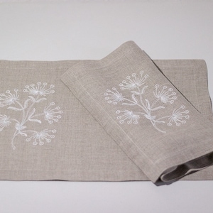 May include: Two beige linen placemats with white embroidered floral designs. The placemats are folded and stacked on top of each other.