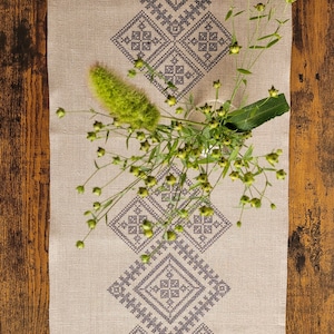 May include: A beige linen table runner with a black embroidered geometric pattern. The runner is decorated with a sprig of green foliage.