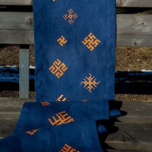 May include: A dark blue linen table runner with embroidered gold geometric patterns. The runner is draped over a wooden bench, showcasing its design and texture. The patterns are evenly spaced and add a decorative element.