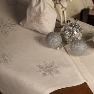 Op de afbeelding: Een feestelijke kerstscène met een witte tafelloper met geborduurde sneeuwvlokken. Een witte cadeauzak met een sneeuwvlokpatroon staat naast zilveren glitterballen en een decoratief paardenbeeldje. De scène heeft een winters thema.