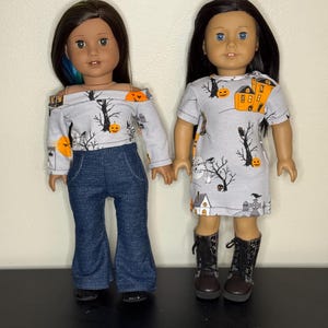 May include: Two dolls wearing Halloween-themed outfits. One doll is wearing a gray off-the-shoulder top with a pumpkin and tree print, paired with blue denim bell-bottom pants. The other doll is wearing a gray dress with a similar print and black boots.